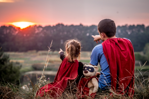 Vader en dochter met superhelden cape wijzen naar de zonsondergang